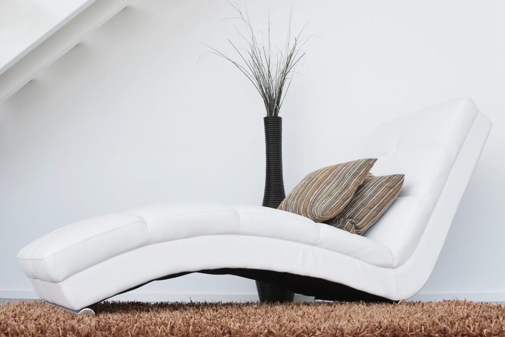 Une chaise longue blanche avec coussins, vase et plante sur tapis